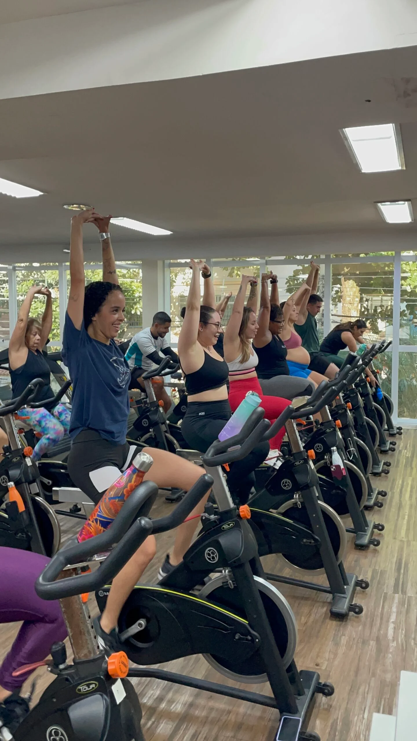 Alunas da MaxFit alongando os braços durante uma aula de bike, com a turma reunida.