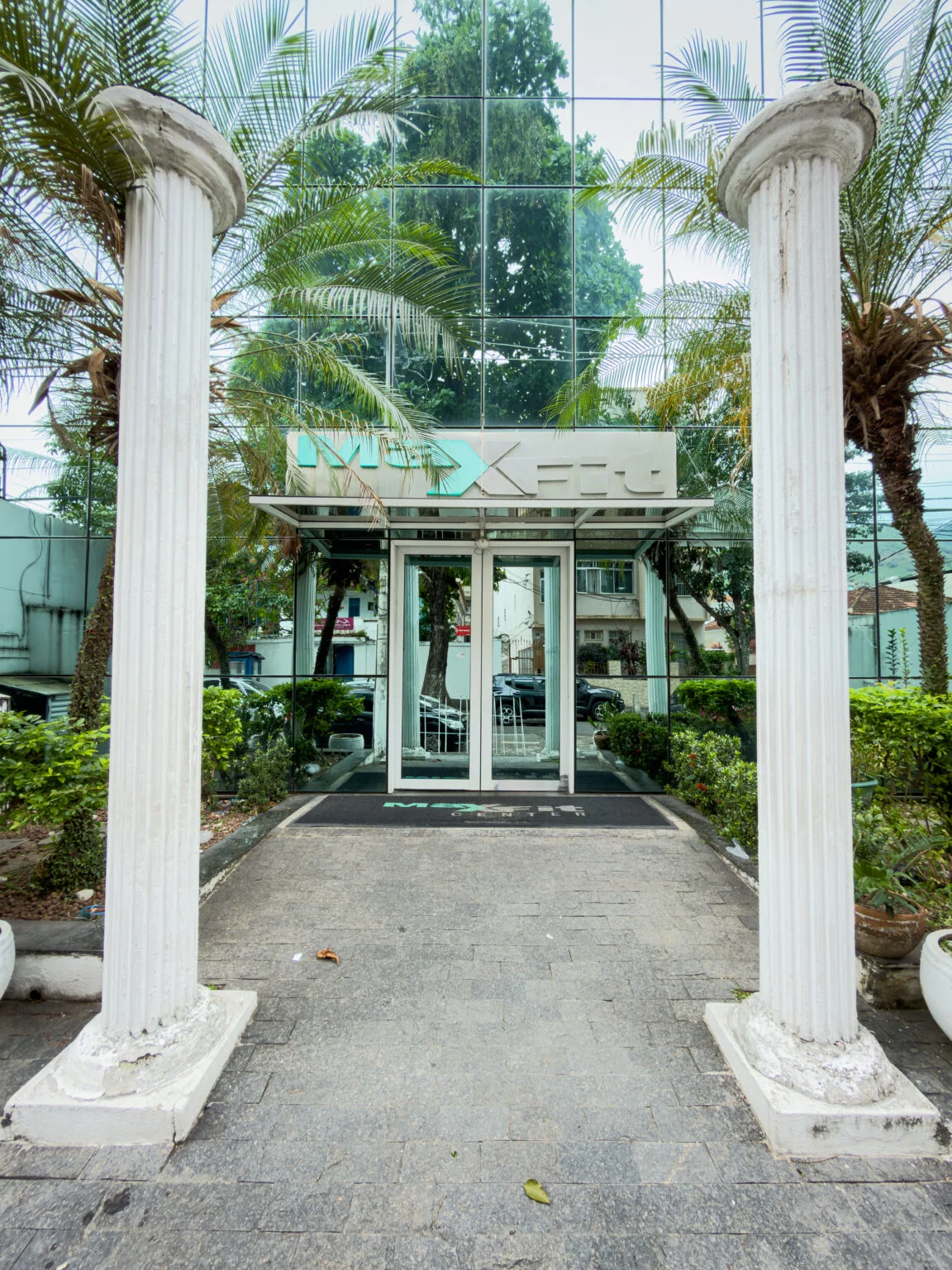 Entrada da MaxFit Center, com fachada de vidro, letreiro da academia e palmeiras.