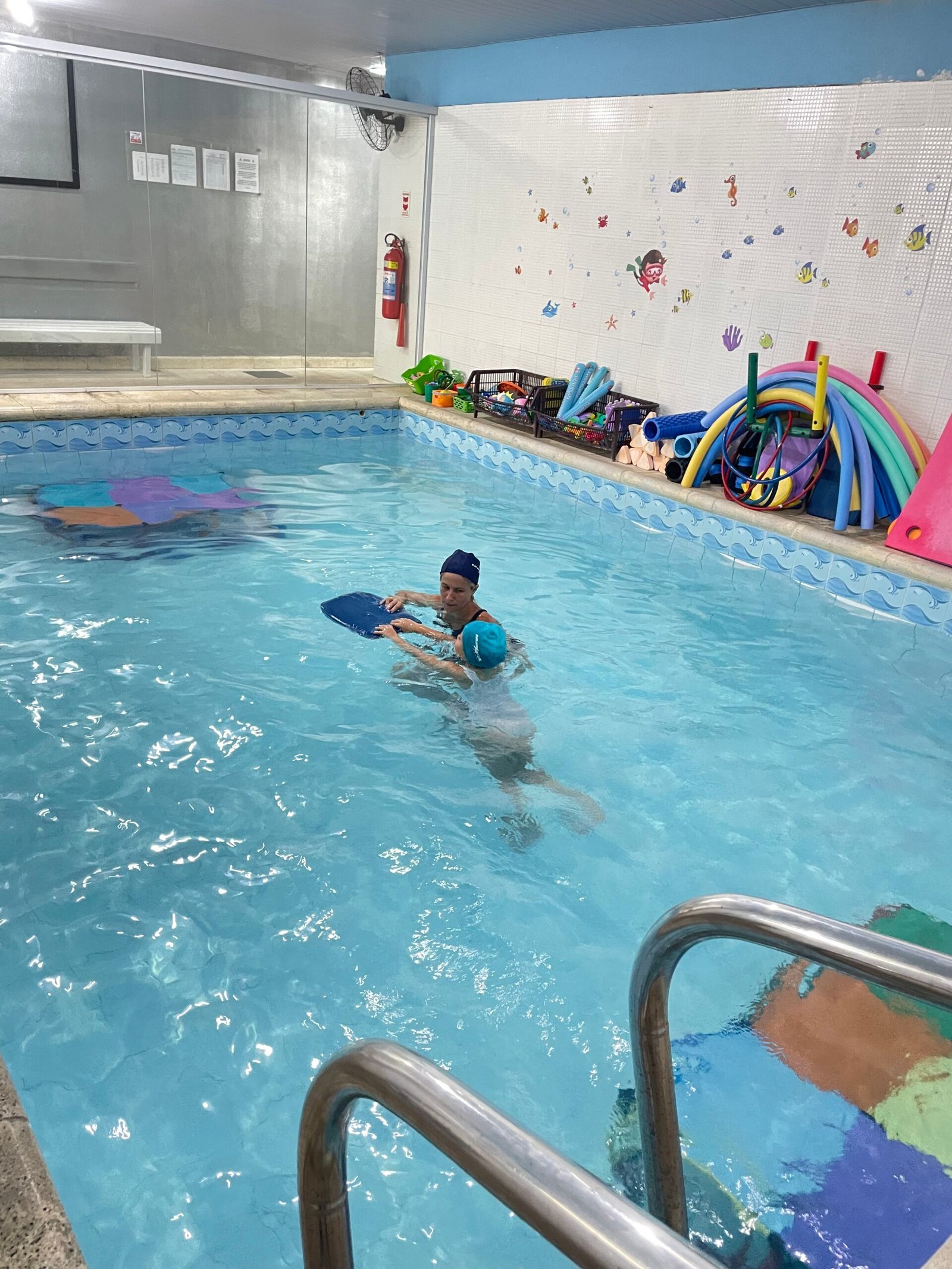 Professora e criança usando uma prancha durante uma aula na piscina infantil da MaxFit.